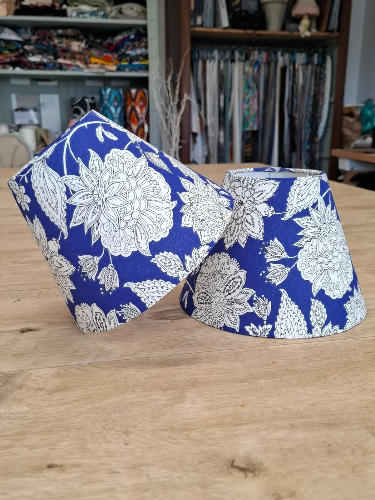Deux abat-jour coniques en tissu bleu à fleurs blanches posés sur la table de l'atelier, rouleaux de tissus en arrière-plan
