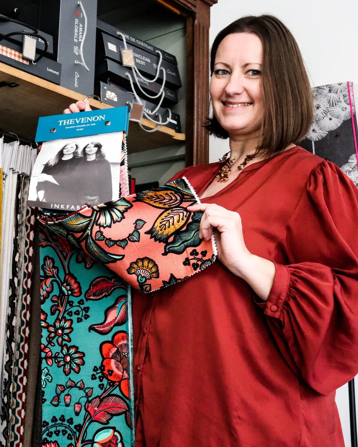 Séverine, souriante, présente un échantillon de tissu fleuri corail devant l'armoire des collections de tissus de l'atelier