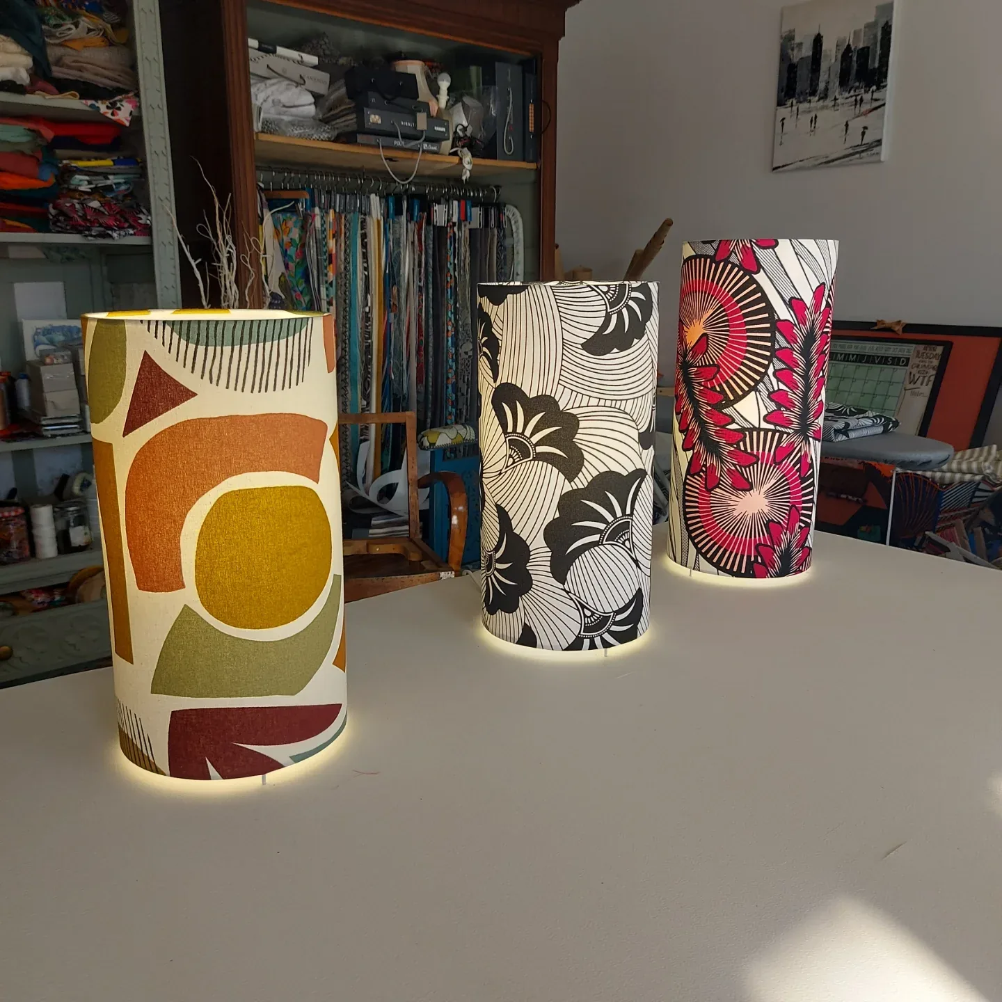 Trois lampes cylindriques réalisées en stage, allumées sur la table de l'atelier devant les rouleaux de tissus