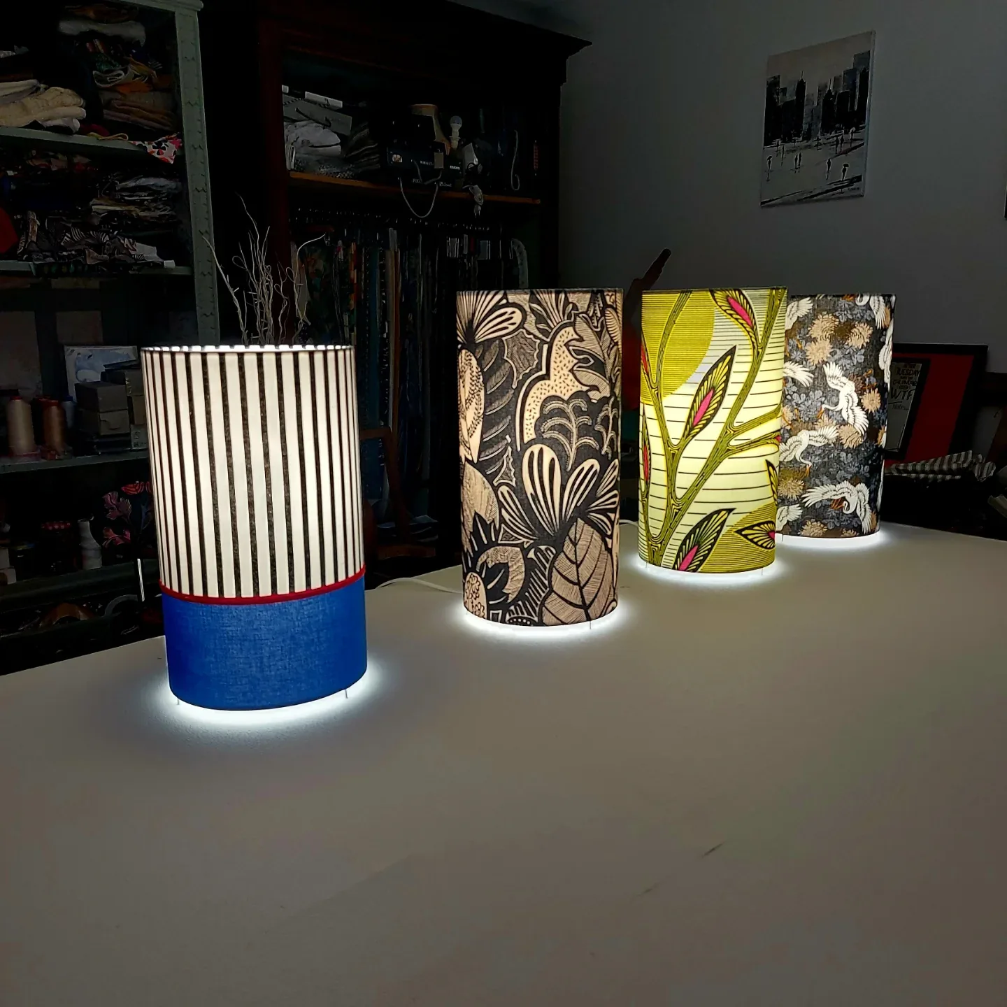 Trois lampes cylindriques en tissu rayé, fleuri et feuillage, allumées dans l'atelier