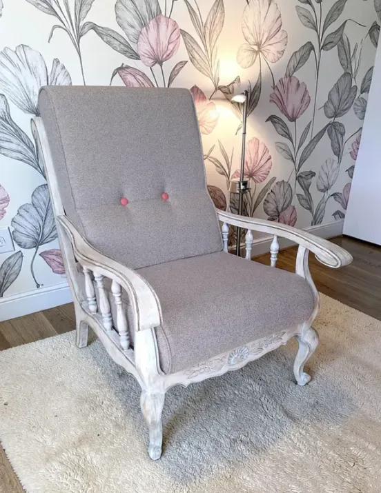 Fauteuil en bois blanchi, tissu gris et boutons corail, devant un papier peint fleuri