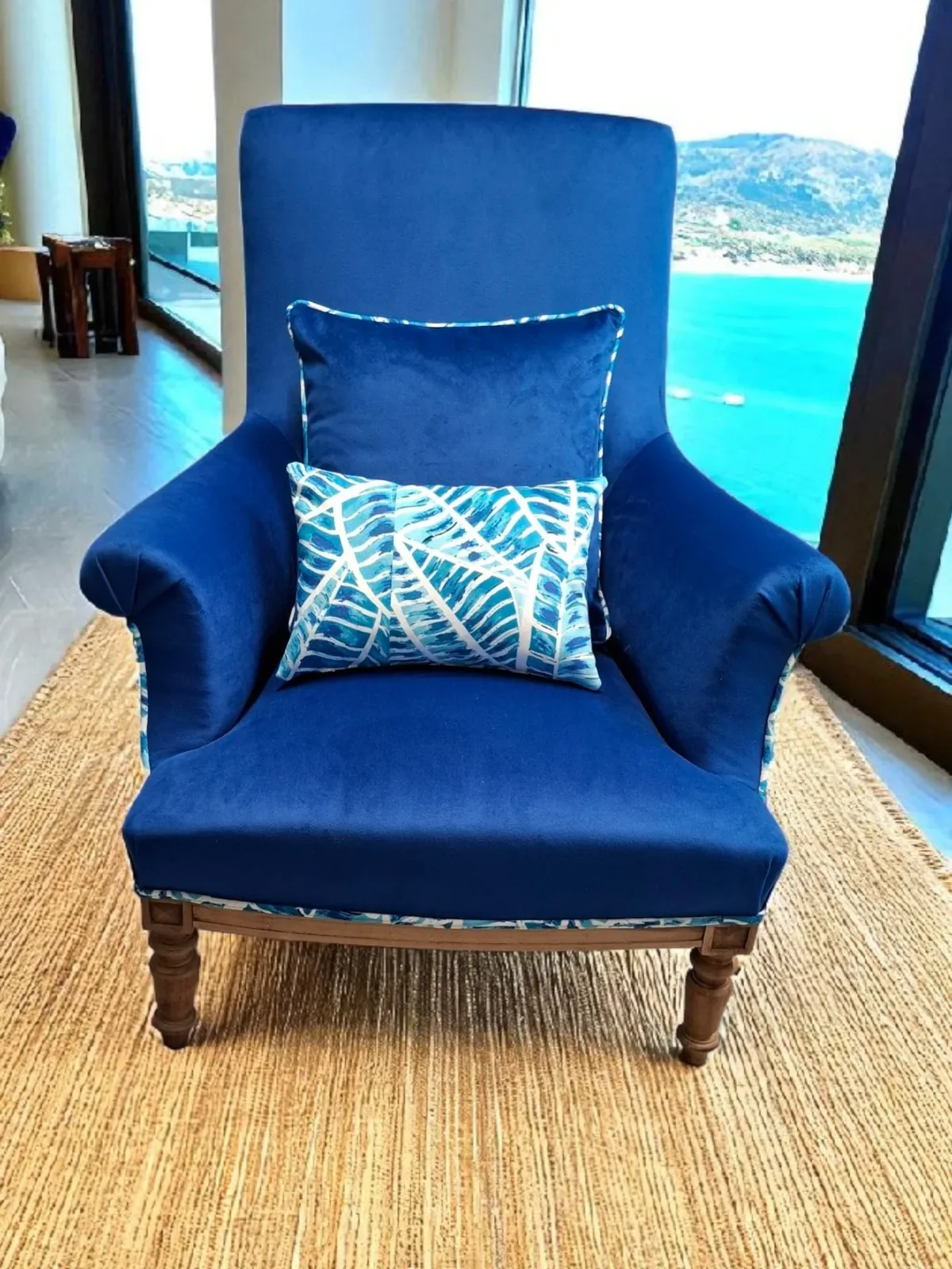 Fauteuil retapissé en velours bleu roi avec coussin à feuillage bleu, devant une baie vitrée donnant sur la mer